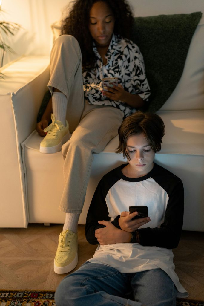 Due adolescenti che usano lo smartphone seduti nello stesso ambiente domestico, simbolo dellenuove dinamiche relazionali nell’era digitale.